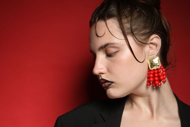 Photo of Beautiful young woman with black lip liner wearing stylish earrings on dark red background, closeup. Space for text
