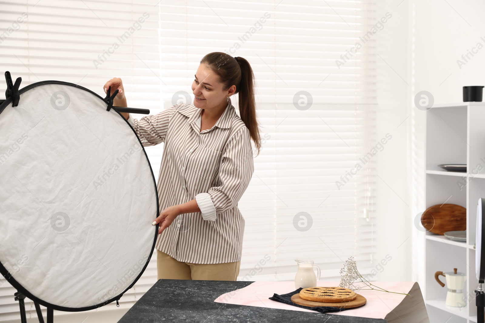 Photo of Woman using reflector to take picture of tasty pie and milk in studio. Professional food photography