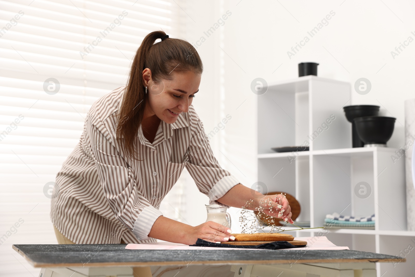Photo of Woman creating composition with tasty pie, milk and floral decor for photoshoot at table in studio. Professional food photography