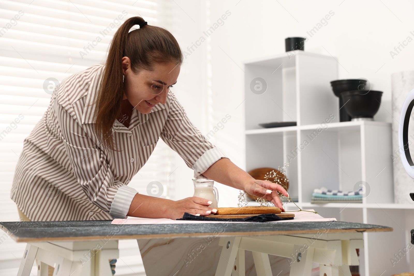 Photo of Woman creating composition with tasty pie, milk and floral decor for photoshoot at table in studio. Professional food photography