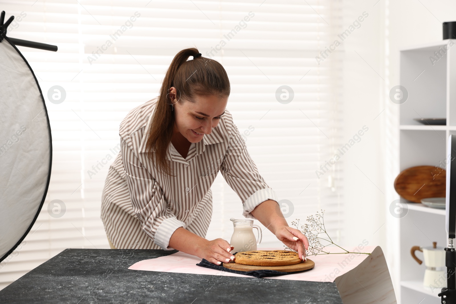 Photo of Woman creating composition with tasty pie, milk and floral decor for photoshoot at table in studio. Professional food photography