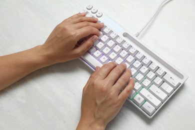 Photo of Woman typing on modern keyboard with RGB lighting at white wooden table, above view
