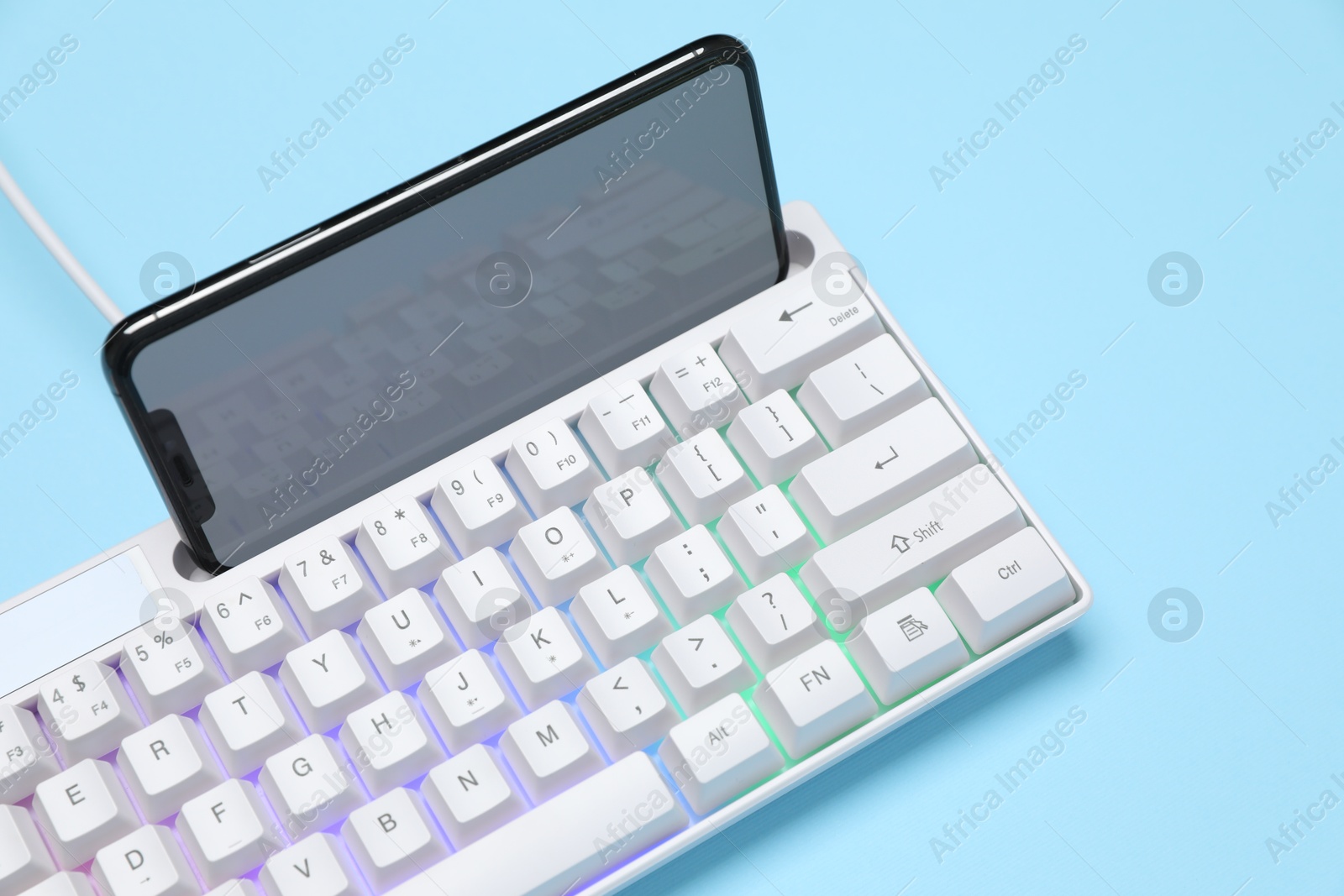 Modern white keyboard with RGB lighting and smartphone on light blue background, closeup Photo of Modern white keyboard with RGB lighting and smartphone on light blue background, closeup