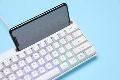 Photo of Modern white keyboard with RGB lighting and smartphone on light blue background, closeup
