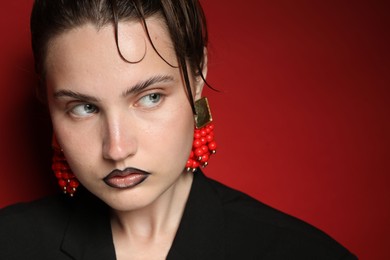 Photo of Beautiful young woman with black lip liner wearing stylish earrings on dark red background, closeup. Space for text