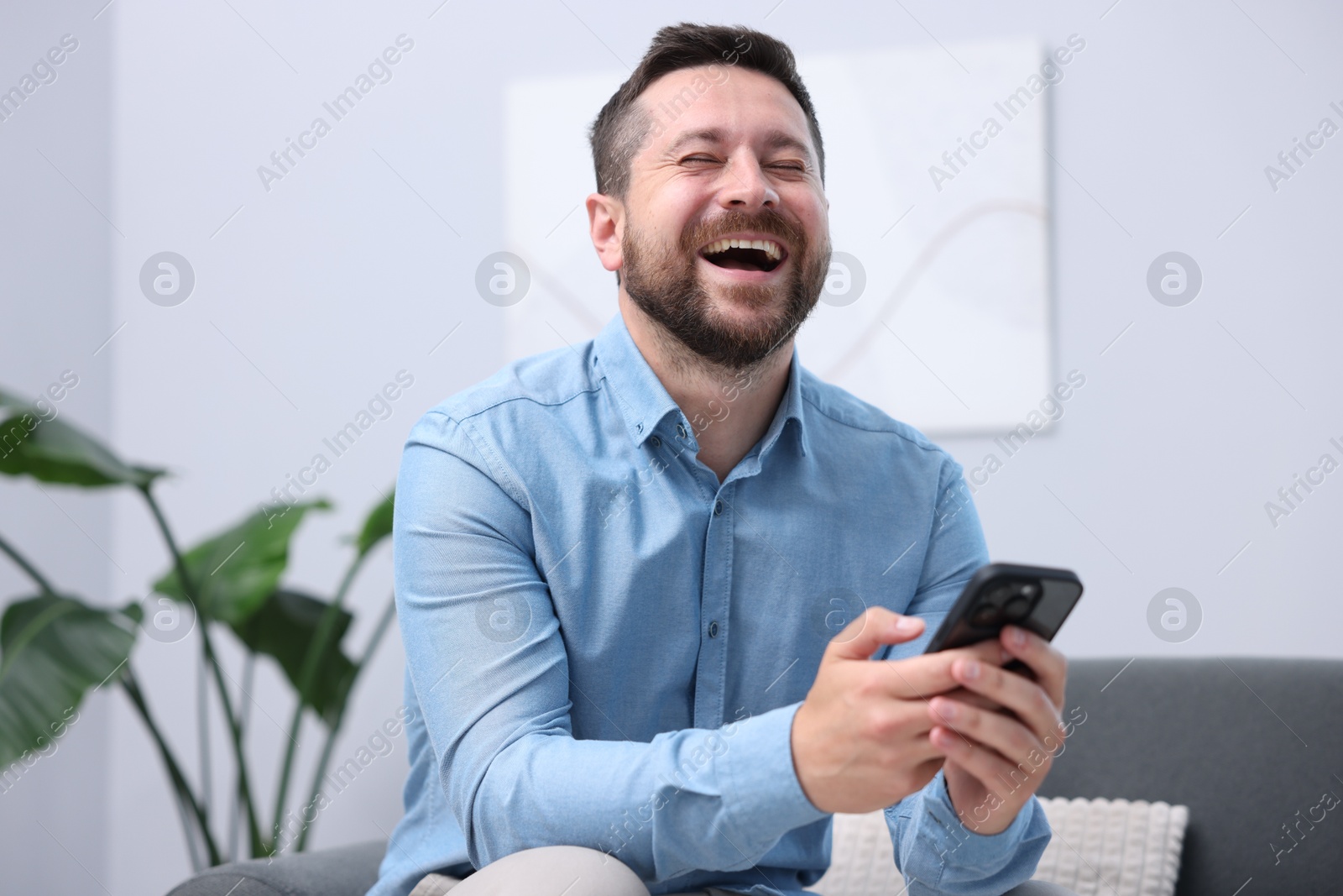 Photo of Man with smartphone laughing on sofa at home