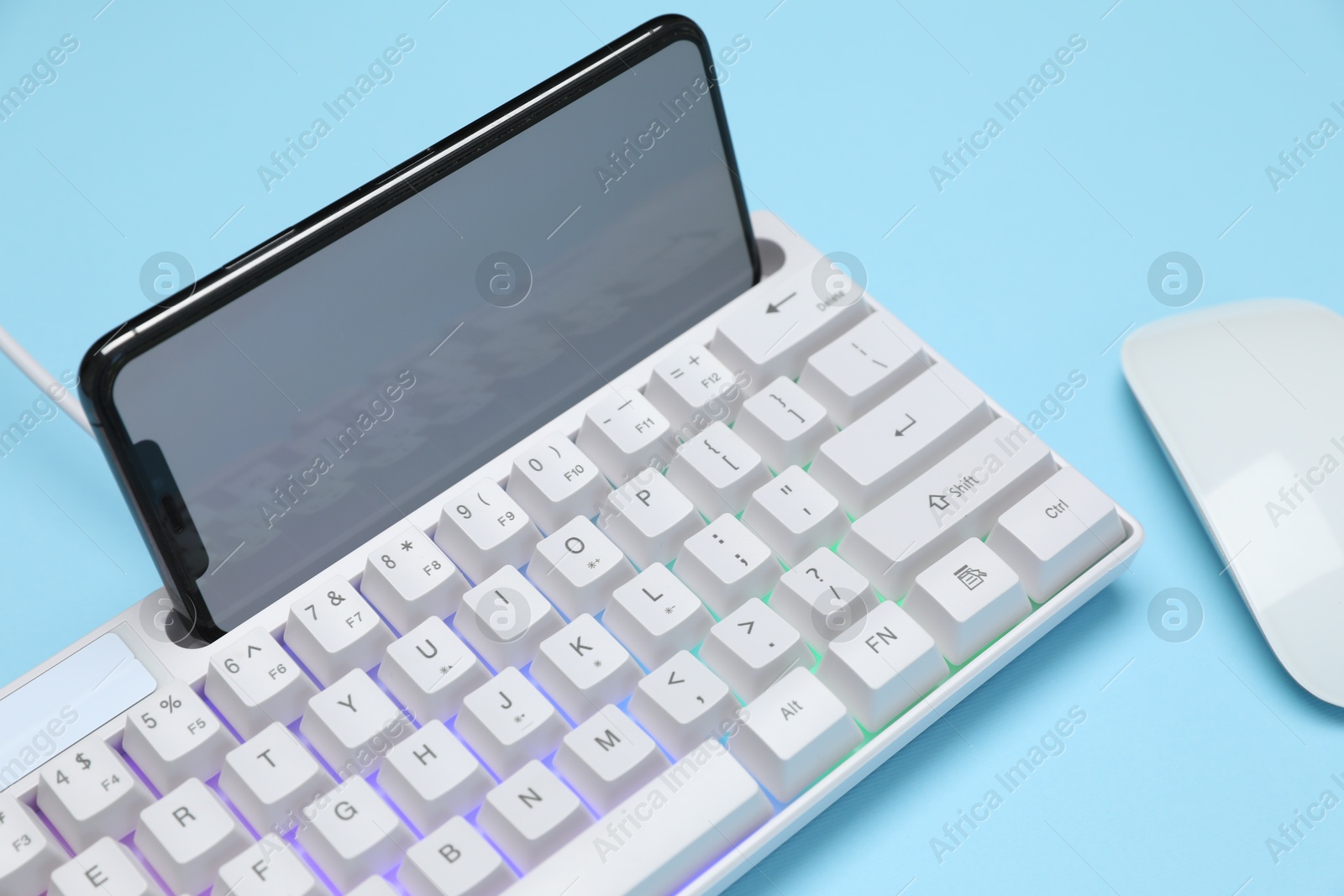 Modern white keyboard with RGB lighting, mouse and smartphone on light blue background, closeup Photo of Modern white keyboard with RGB lighting, mouse and smartphone on light blue background, closeup
