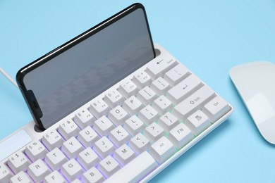 Photo of Modern white keyboard with RGB lighting, mouse and smartphone on light blue background, closeup