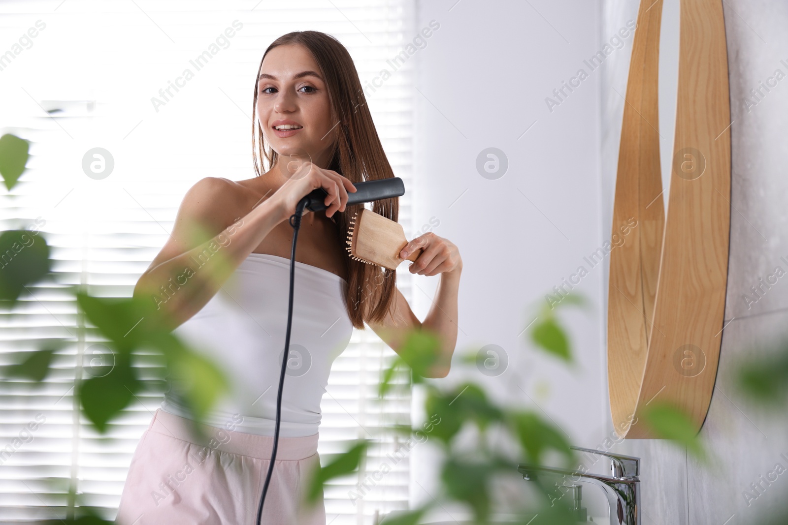 Photo of Getting ready. Young woman brushing her hair and using straightener at home