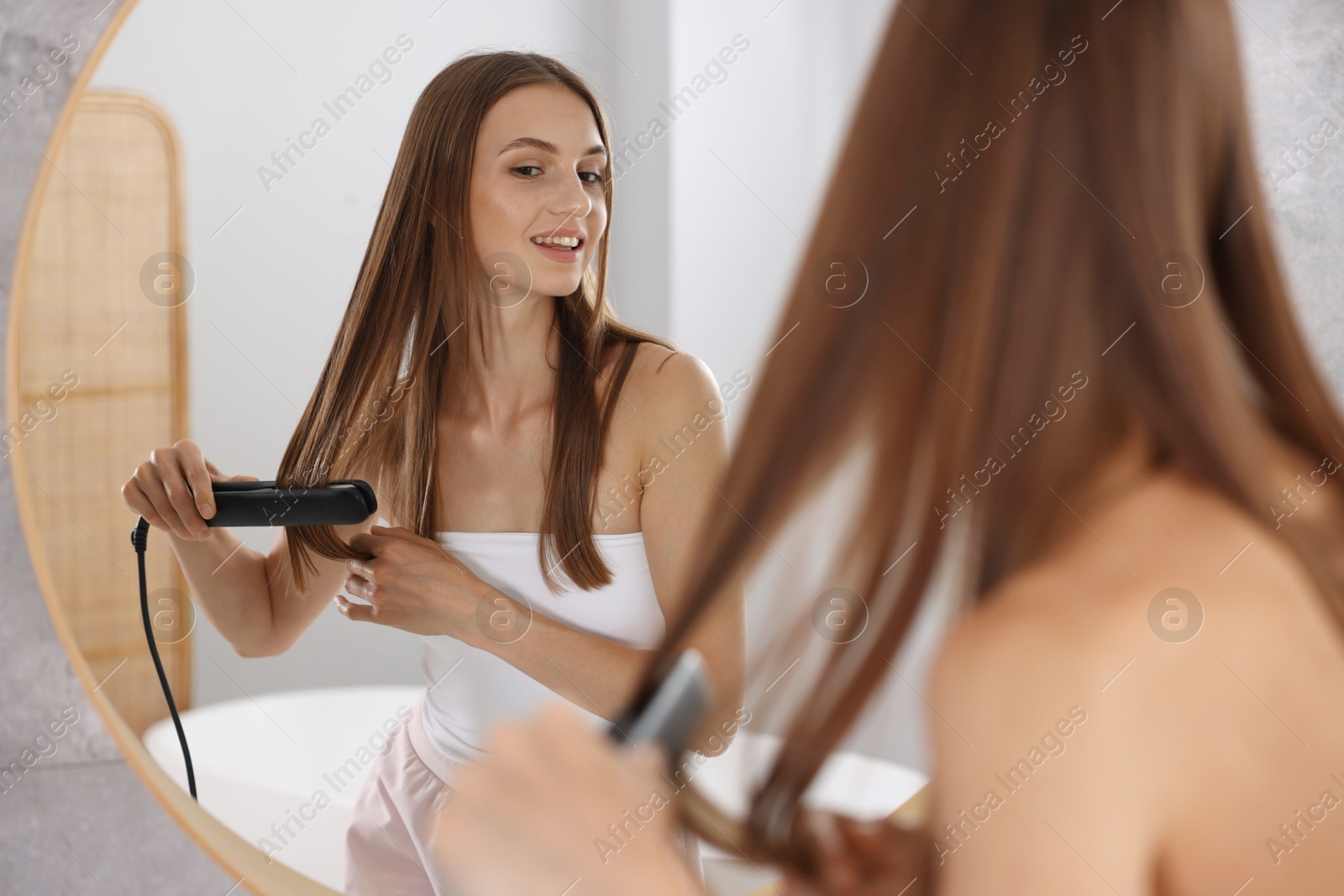 Photo of Getting ready. Young woman using hair straightener at home