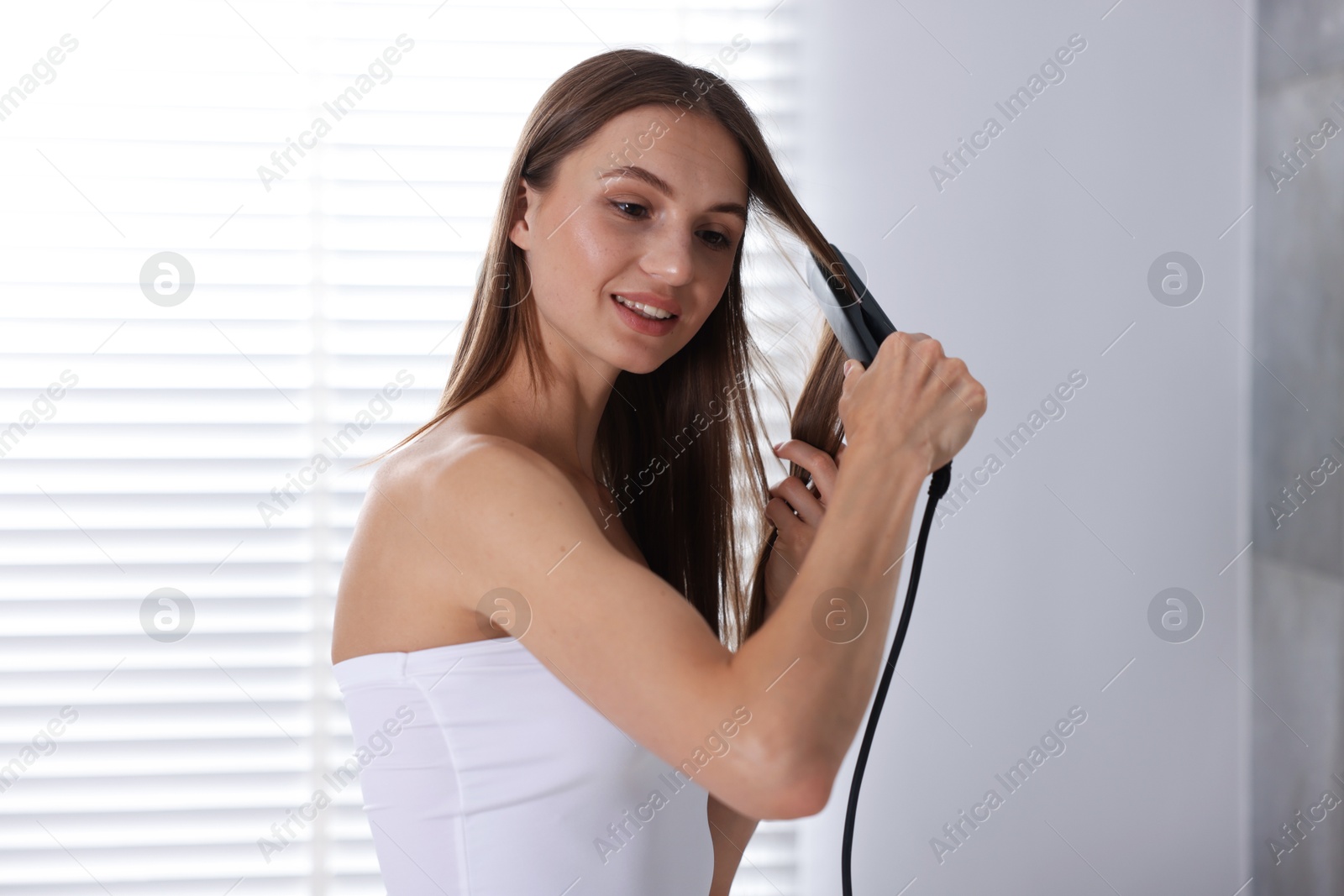 Photo of Getting ready. Young woman using hair straightener at home