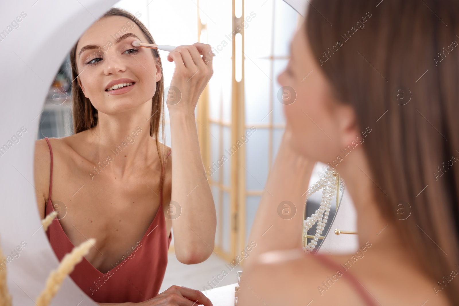 Photo of Getting ready. Young woman applying makeup near mirror at vanity indoors