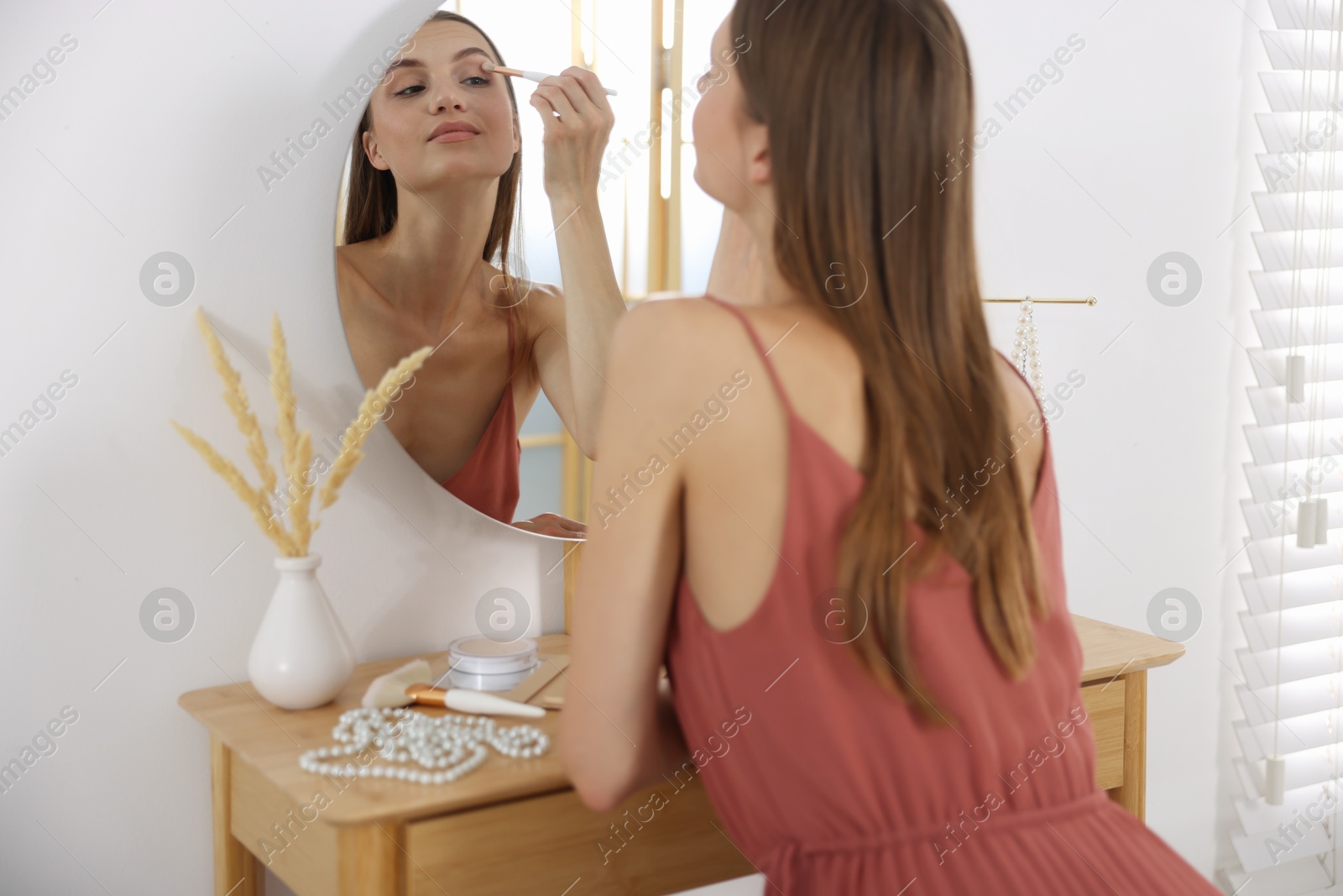 Photo of Getting ready. Young woman applying makeup near mirror at vanity indoors