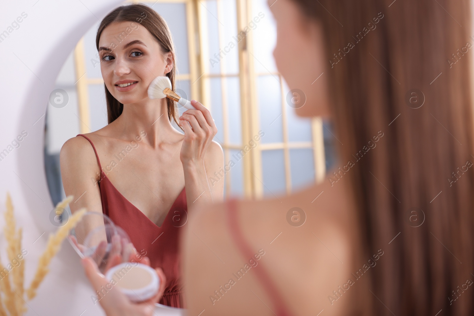 Photo of Getting ready. Young woman applying makeup near mirror at vanity indoors
