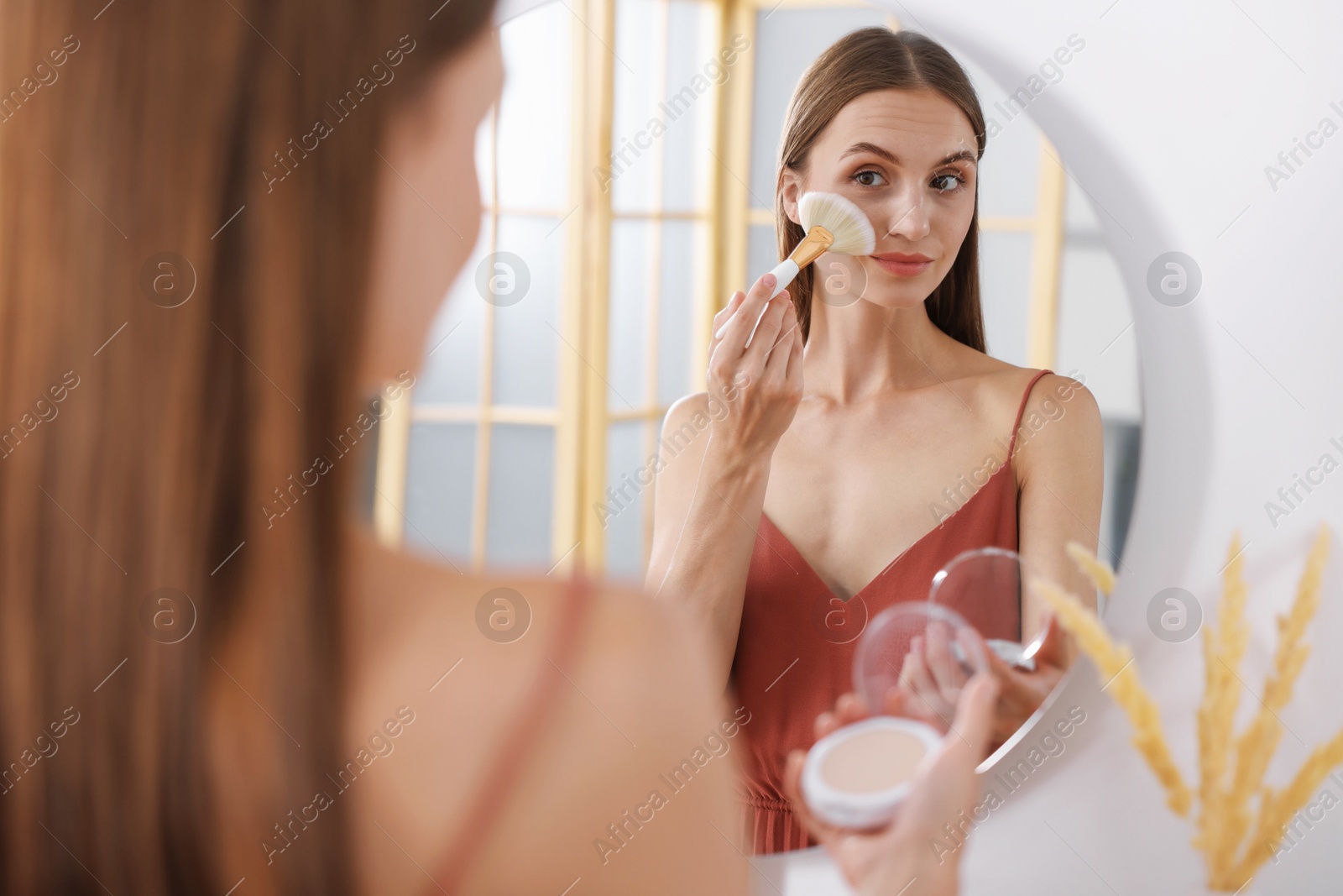 Photo of Getting ready. Young woman applying makeup near mirror at vanity indoors