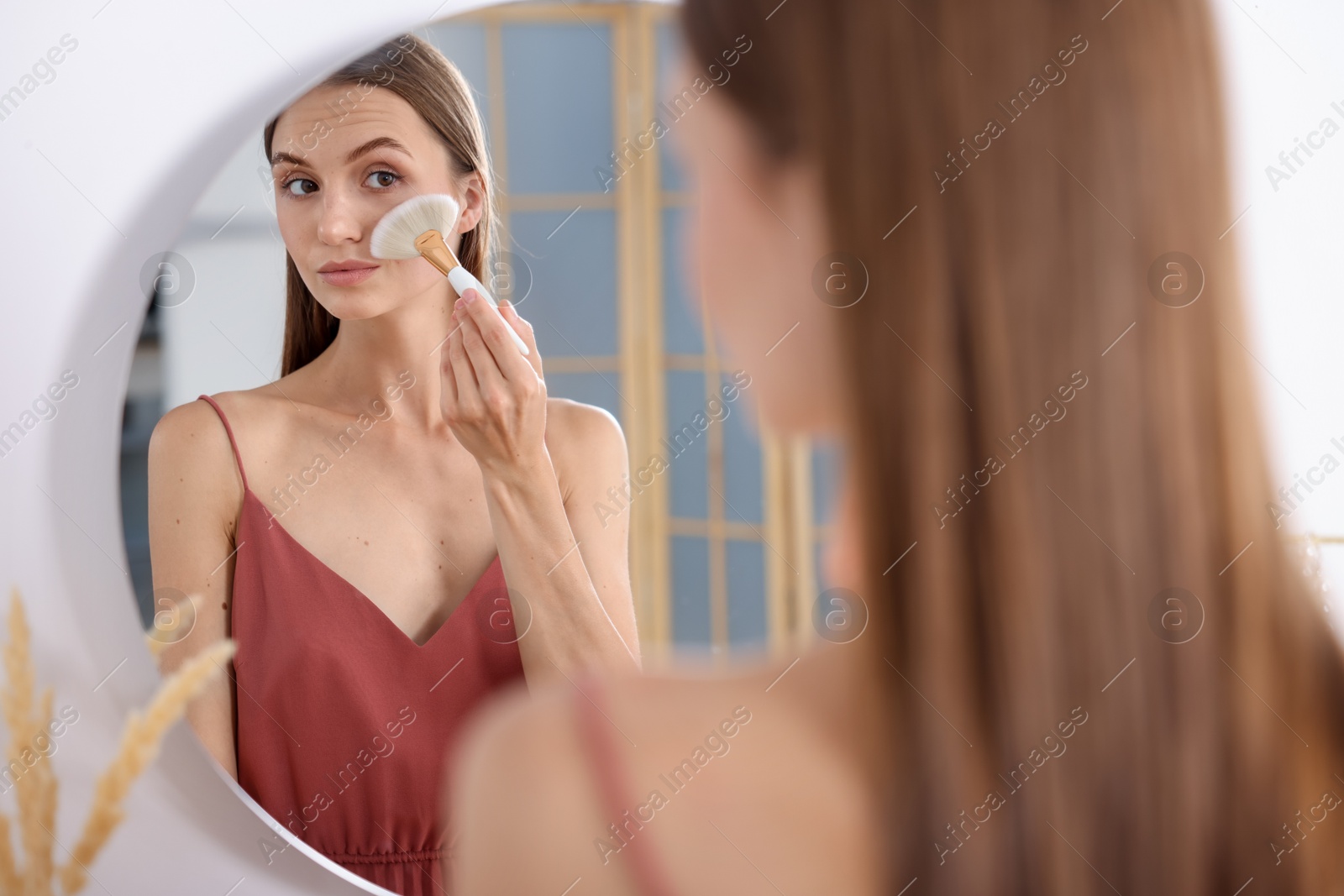 Photo of Getting ready. Young woman applying makeup near mirror at vanity indoors