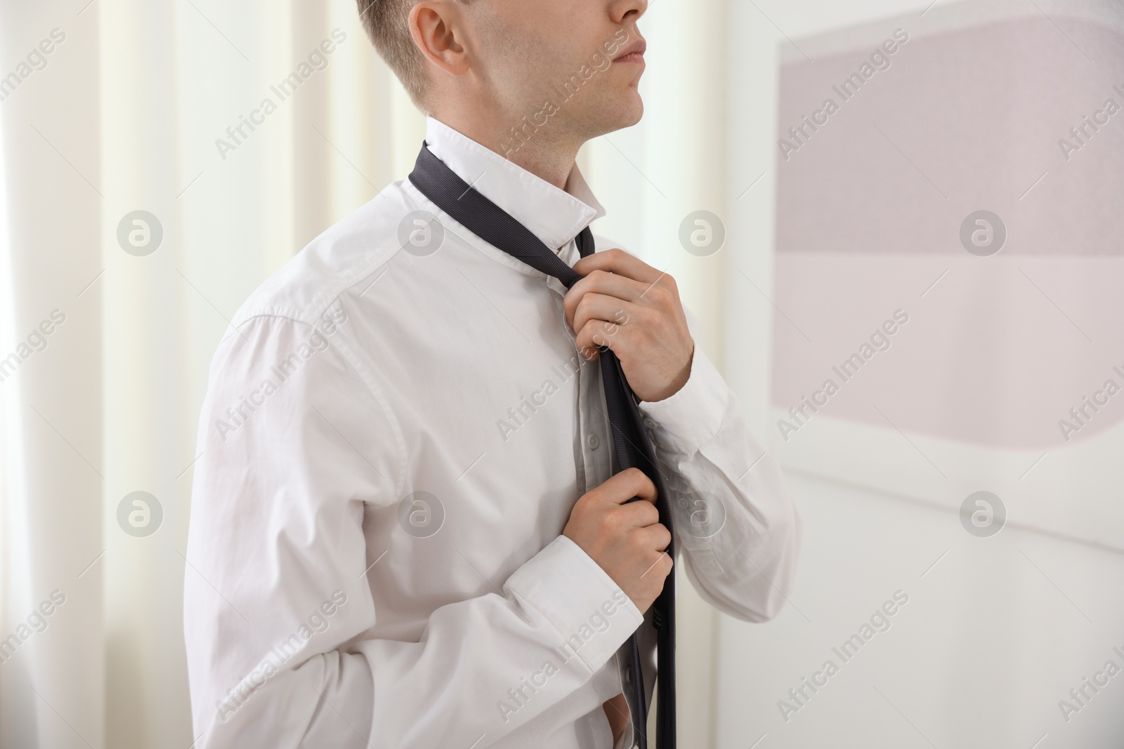 Photo of Getting ready. Young man tying stylish necktie at home, closeup