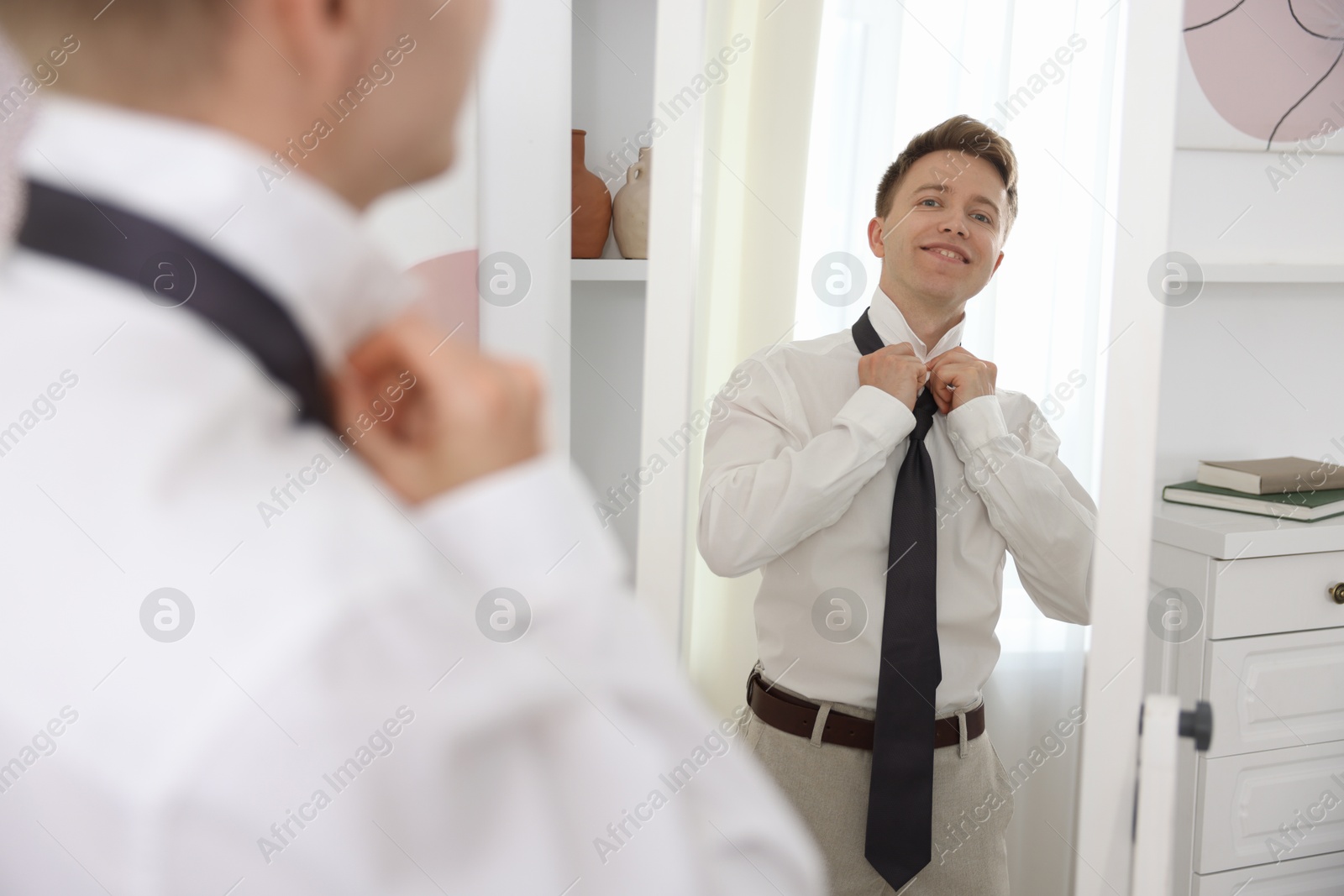 Photo of Getting ready. Young man tying stylish necktie near mirror at home