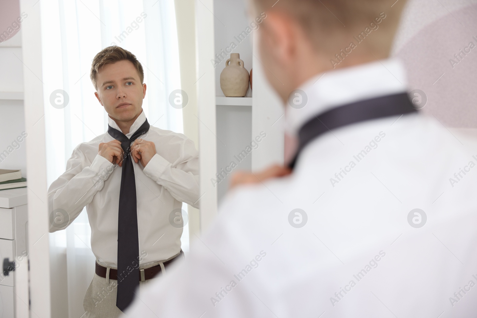 Photo of Getting ready. Young man tying stylish necktie near mirror at home