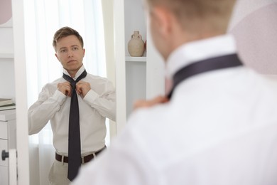 Photo of Getting ready. Young man tying stylish necktie near mirror at home