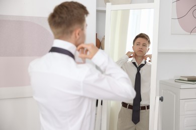 Photo of Getting ready. Young man tying stylish necktie near mirror at home