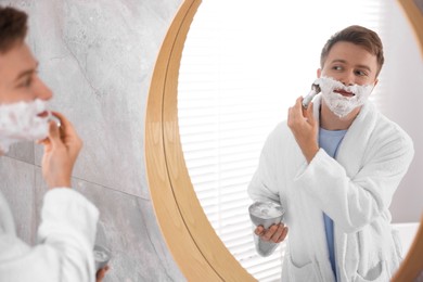 Photo of Getting ready. Young man shaving with cream near mirror at home
