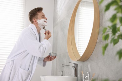 Photo of Getting ready. Young man shaving with cream near mirror at home