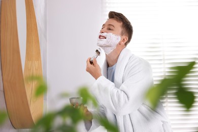 Photo of Getting ready. Young man shaving with cream near mirror at home
