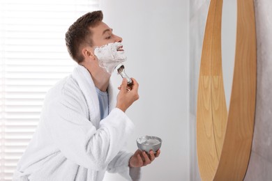 Photo of Getting ready. Young man shaving with cream near mirror at home