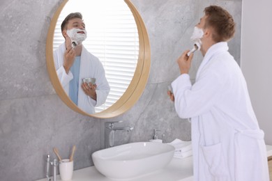 Photo of Getting ready. Young man shaving with cream near mirror at home