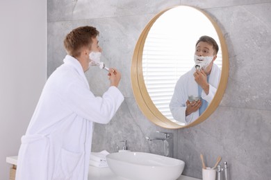 Photo of Getting ready. Young man shaving with cream near mirror at home