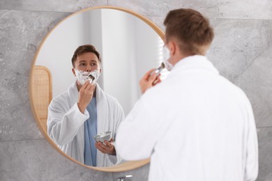 Photo of Getting ready. Young man shaving with cream near mirror at home
