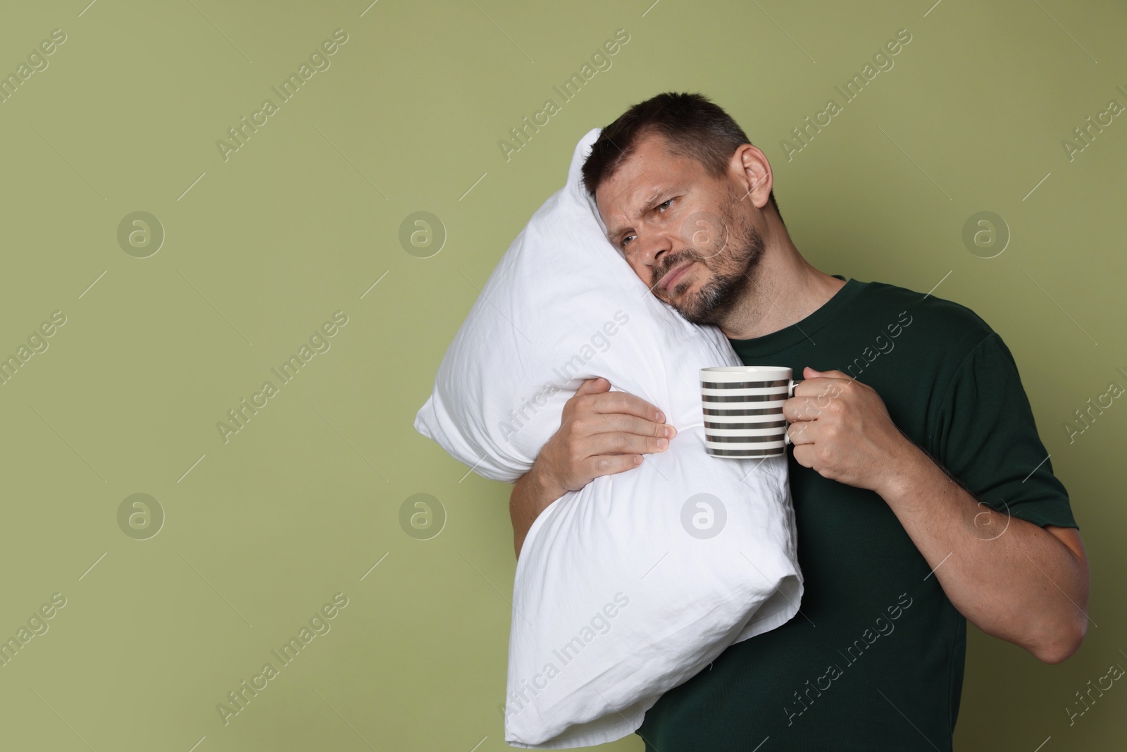 Photo of Man with pillow and cup of drink suffering from hangover on olive background, space for text