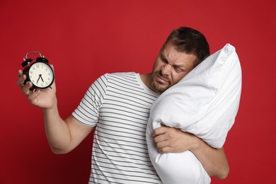 Photo of Man with pillow and alarm clock suffering from hangover on red background