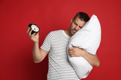 Photo of Man with pillow and alarm clock suffering from hangover on red background
