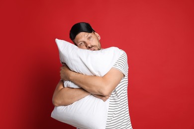Photo of Man with pillow and sleep mask suffering from hangover on red background
