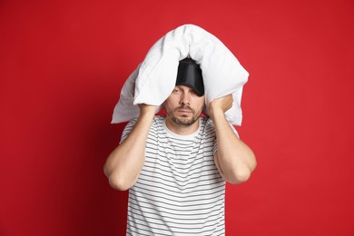 Photo of Man with pillow and sleep mask suffering from hangover on red background