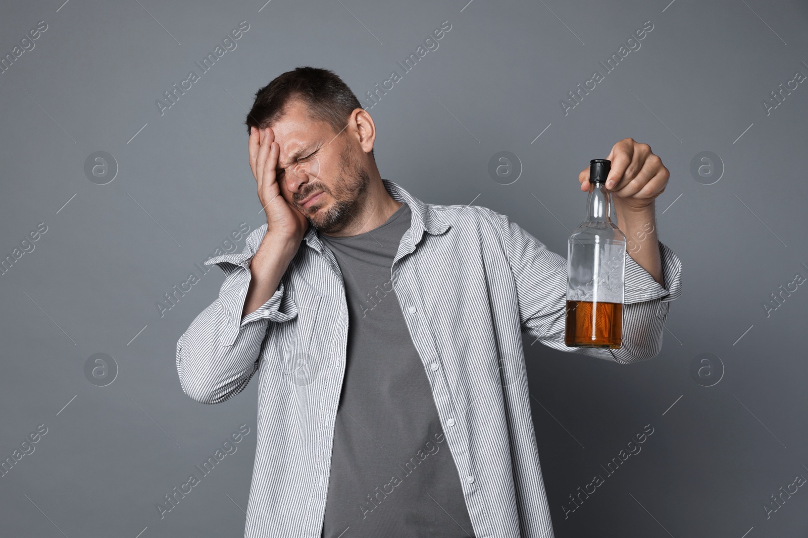 Photo of Man with bottle of whiskey suffering from hangover on grey background