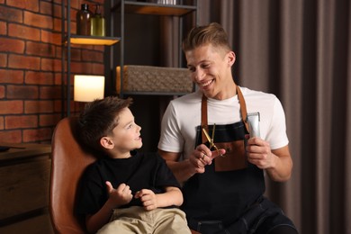 Photo of Professional barber with scissors, trimmer and little boy in barbershop