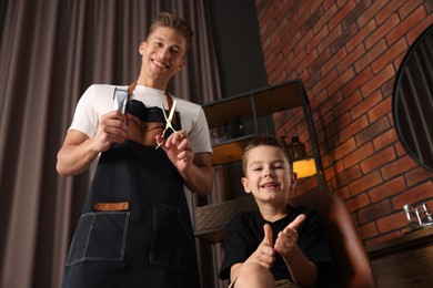 Photo of Professional barber with scissors, trimmer and little boy in barbershop, low angle view