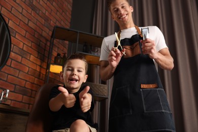 Photo of Professional barber with scissors, trimmer and little boy in barbershop, low angle view