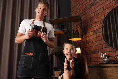 Photo of Professional barber with scissors, trimmer and little boy in barbershop, low angle view