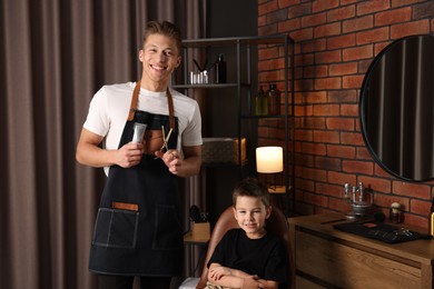 Photo of Professional barber with scissors, trimmer and little boy in barbershop