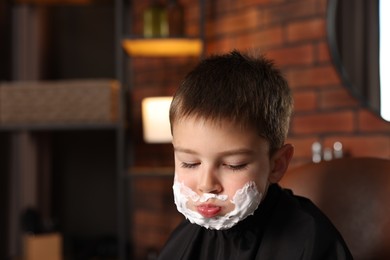 Photo of Little boy with shaving foam on his face in barbershop, space for text