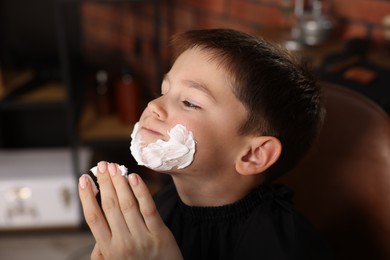 Photo of Professional barber applying shaving foam onto boy's face in barbershop, closeup