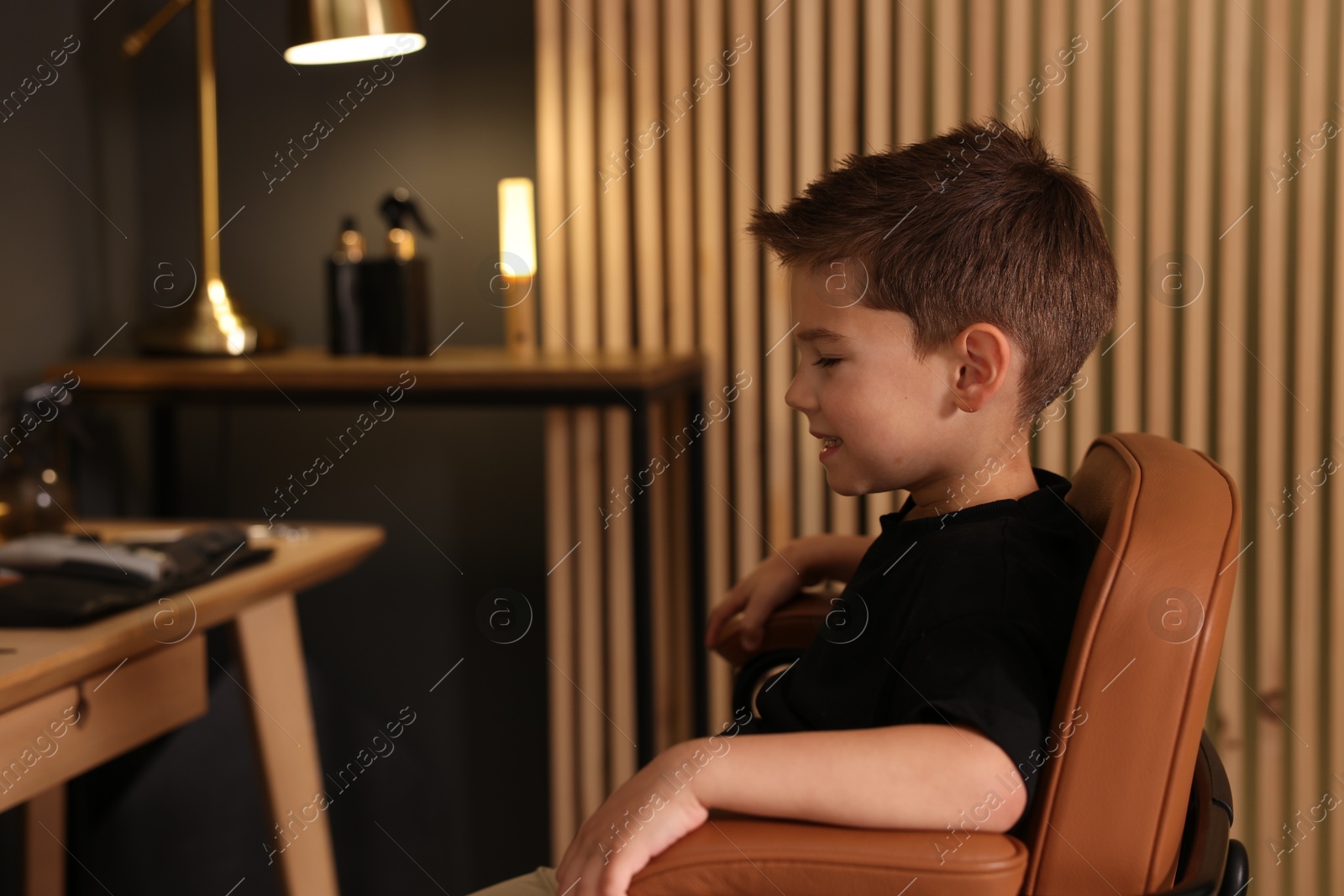 Photo of Cute little boy sitting in chair in barbershop, space for text