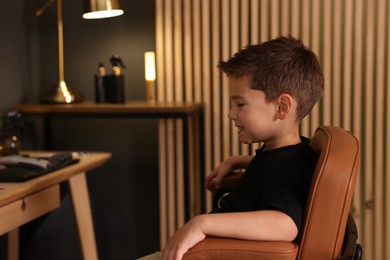 Photo of Cute little boy sitting in chair in barbershop, space for text
