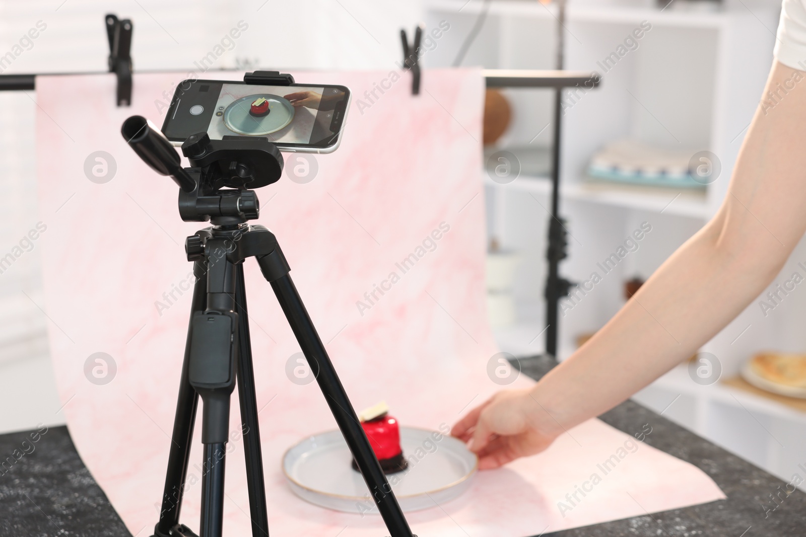 Photo of Woman creating composition with tasty dessert for photoshoot at table in studio, focus on smartphone and tripod. Professional food photography