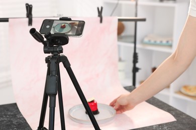 Photo of Woman creating composition with tasty dessert for photoshoot at table in studio, focus on smartphone and tripod. Professional food photography