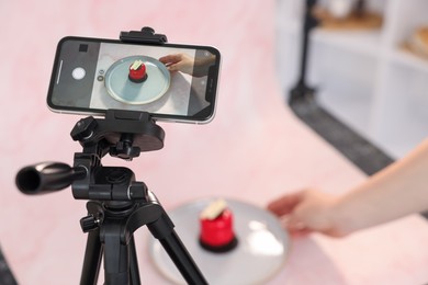Photo of Woman creating composition with tasty dessert for photoshoot at table in studio, focus on smartphone and tripod. Professional food photography